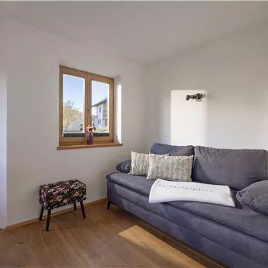 A cozy living room with a gray sofa and bright windows. The wooden floor and neutral colors create an inviting atmosphere.