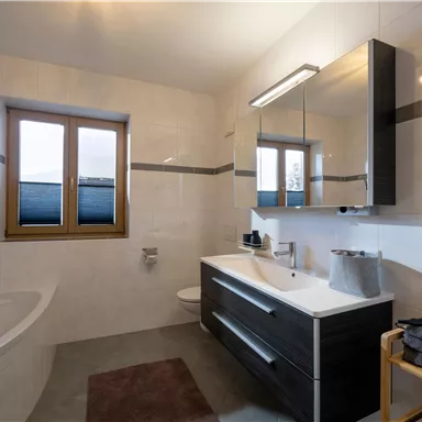 A modern bathroom with a large mirror and a sink. The walls are bright, and there is a window that lets in daylight.