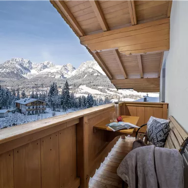 A balcony with wooden cladding, featuring a cozy seating area. In the background, snow-covered mountains rise against a clear sky.