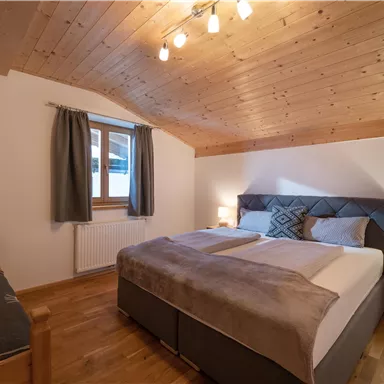 A cozy bedroom with a wooden ceiling and warm light. There is a large bed and a comfortable armchair in the corner.