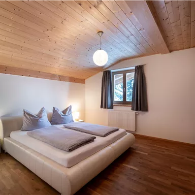 A cozy bedroom with two beds and a wooden ceiling. The room is bright and modernly furnished, with windows and a lamp.