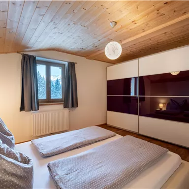 A modern bedroom with a wooden ceiling and a large window. The beds are equipped with light pillows and blankets, and a spacious wardrobe is in the room.