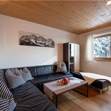 A modern living room with a black sofa and cozy cushions. Large windows offer a view of a snow-covered landscape.