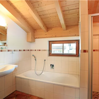 A modern bathroom with a bathtub and a separate shower. The wooden ceiling and the light tiles create a cozy atmosphere.