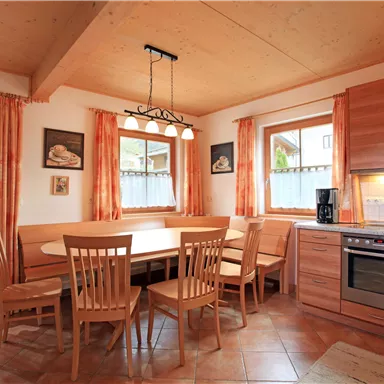 A cozy kitchen with a large dining table and wooden furniture. Bright windows with curtains create a friendly atmosphere.