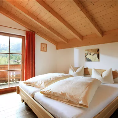 A cozy bedroom with wooden ceilings and a double bed. Large windows offer a view of the mountains and the balcony.
