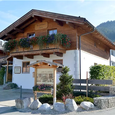 A traditional wooden house with a balcony and blooming plants. In the background, gentle hills can be seen.