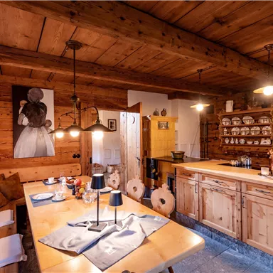 A cozy wooden house kitchen with a large dining table and a rustic counter area. The walls are decorated with wooden paneling and kitchen utensils.