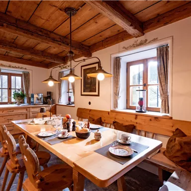 A cozy kitchen with wooden furniture and a large table. The table is set and the light falls softly through the windows.