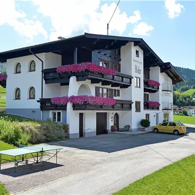 A quiet country house with balconies full of blooming flowers. In the foreground, there is a table and a yellow car on the paved driveway.