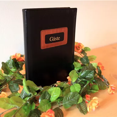 Ein Gästebuch steht auf einem Tisch, umgeben von künstlichen Blumen und grünen Blättern. Die Atmosphäre ist einladend und freundlich.