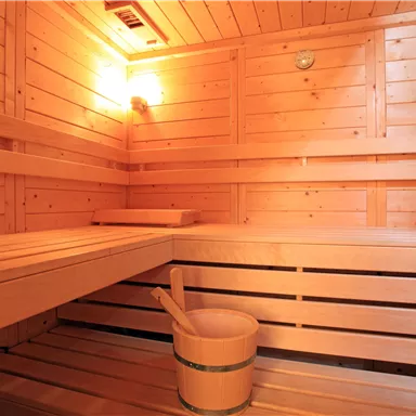 Eine gemütliche Sauna mit Holzwänden und Bänken. In der Mitte steht ein Holz-Eimer mit einem Schöpflöffel.