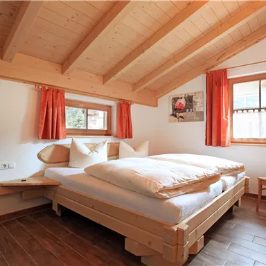 Ein gemütliches Schlafzimmer mit einem Holzdesign und einem schrägen Dach. Es gibt ein Doppelbett, zwei Fenster mit roten Vorhängen und einen Stuhl aus Holz.