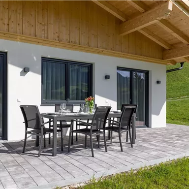 A modern terrace with a dining table and chairs. In the background, a green hillside and large windows are visible.