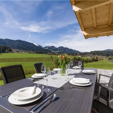 An elegant outdoor dining area with views of green meadows and mountains. The table is beautifully set with dishes and flowers.