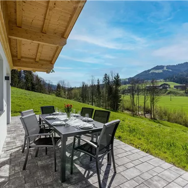 A modern terrace with a dining table and chairs. In the background, green meadows and mountains stretch under a clear blue sky.