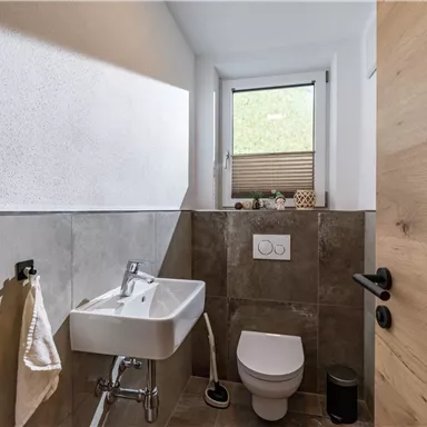 A modern bathroom with gray tiles and a small window. There is a sink and a toilet, as well as natural wood elements.