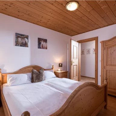 A cozy bedroom with a large double bed and wooden ceiling. The room features wooden furniture and decorative pictures on the walls.