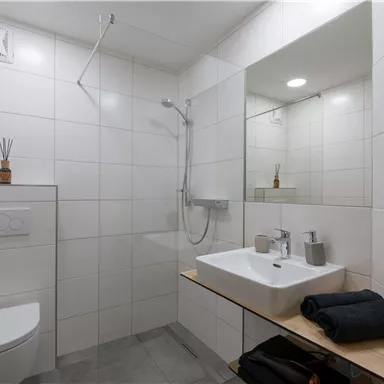 A modern bathroom with white tiles and an open shower. On the wooden shelf are towels and toiletries.