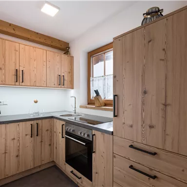 A modern kitchen with wooden furniture and a clear countertop. It features a stove, a sink, and large windows that let in plenty of light.