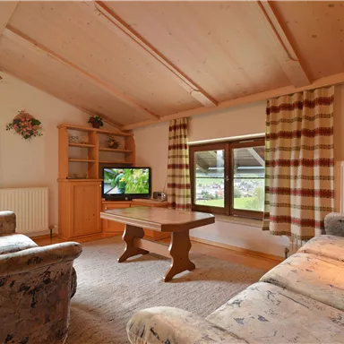 Ein gemütliches Wohnzimmer mit Holzdecke und großen Fenstern. Es gibt einen Fernseher, einen Couchtisch und komfortable Möbel.