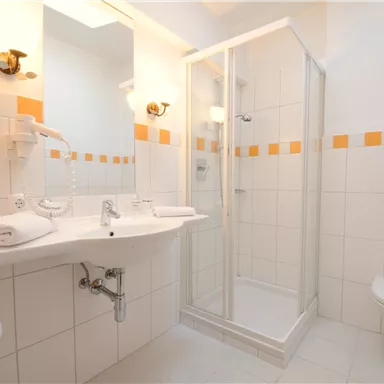 A modern bathroom with a shower and a sink. The walls are light-tiled and decorated with orange accents.