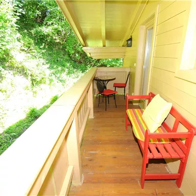 A cozy balcony with red seating and a small table. In the background, lush green foliage can be seen.