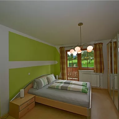 A modern bedroom with green walls and large windows. There is a double bed, a nightstand, and a wardrobe.