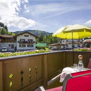 Ein gemütlicher Balkon mit einem roten Stuhl und einem gelben Sonnenschirm. Im Hintergrund sind schöne Berge und traditionelle österreichische Häuser zu sehen.