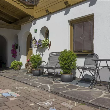 Ein einladender Außenbereich mit Sitzgelegenheiten und blühenden Pflanzen. Die helle Fassade und der gepflasterte Weg schaffen eine gemütliche Atmosphäre.