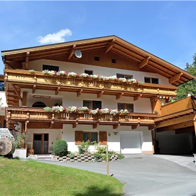 A lovely chalet in alpine style with wooden balconies adorned with carvings and beautiful flowers. Surrounded by trees and a green meadow.