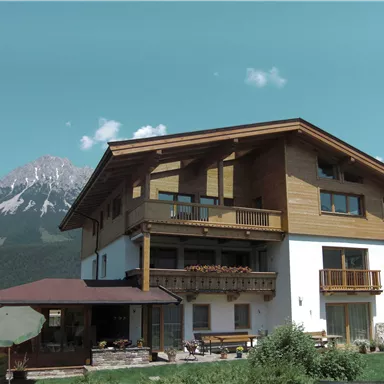 A modern house with wooden elements and balconies in a mountainous setting. In the background, snow-capped mountains and a clear sky can be seen.