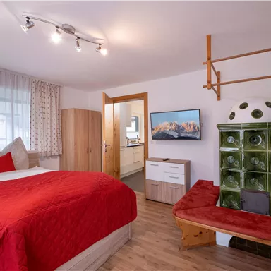Ein gemütliches Schlafzimmer mit einem roten Bett und modernen Möbeln. Teils Landhausstil mit einem Ofen und einem großen Fenster.