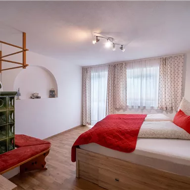 Ein gemütliches Schlafzimmer mit einem großen Bett und roten Kissen. Der Raum hat einen Holzfußboden und helle Fenster mit Vorhängen.