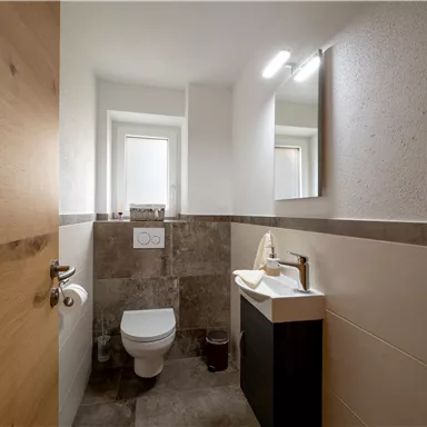 Ein modernes Badezimmer mit einem weißen Waschbecken und einer Toilette. Es gibt Belegaungen in hellen und dunklen Farbtönen sowie ein Fenster für natürliches Licht.