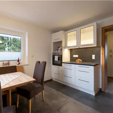 Eine moderne Küche mit hellen Schränken und einem Esstisch. Das Zimmer ist gut beleuchtet und hat große Fenster.