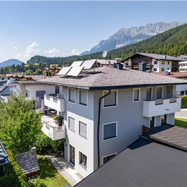 Ein modernes Haus mit mehreren Etagen und Solarpanelen. Im Hintergrund sind grüne Berge und eine ruhige Wohngegend zu sehen.