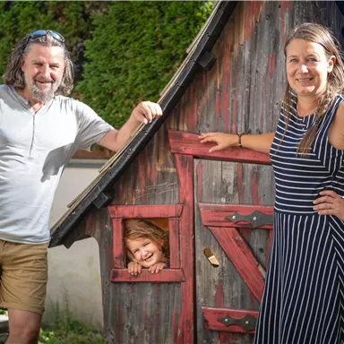 A happy couple stands next to a small wooden house. A child looks cheerfully out of the window of the house.
