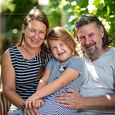 Eine glückliche Familie sitzt im Garten. Die Mutter, der Vater und das Kind lächeln fröhlich in die Kamera.