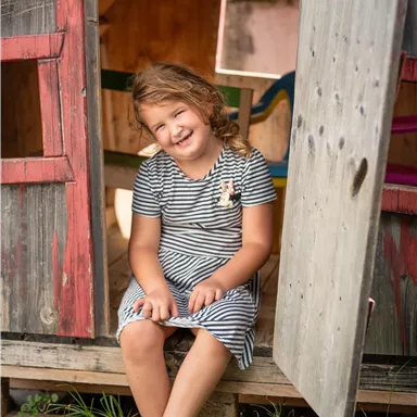 A happy girl is sitting in a small playhouse. She is wearing a striped dress and is smiling.