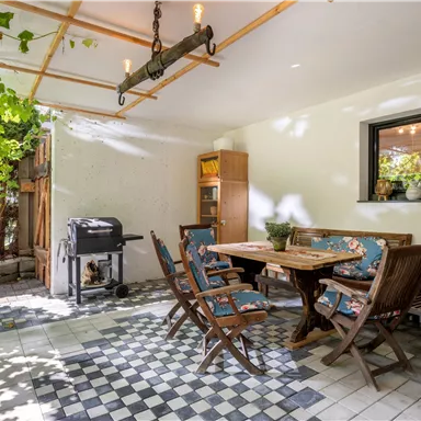 Eine gemütliche Terrasse mit einem Esstisch und Stühlen aus Holz. Im Hintergrund stehen ein Grill und eine Pflanze, die Schatten bietet.