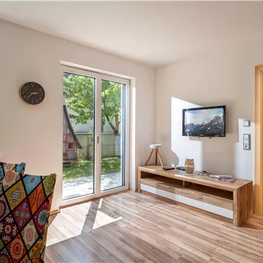 A modern living room with large windows that let in a lot of daylight. There is a television, a wooden cabinet, and a colorful combination of chairs.