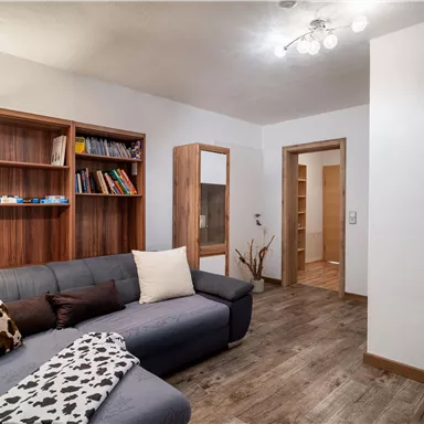 Ein gemütliches Wohnzimmer mit einem grauen Sofa und Regalen. Der Raum hat warmes Licht und Holzkolorierungen, die eine einladende Atmosphäre schaffen.