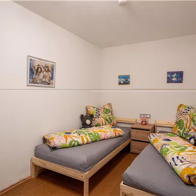 A simple bedroom with two beds and colorful bedding. There are some pictures on the wall and a small nightstand between the beds.