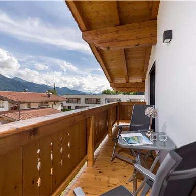 Ein gemütlicher Balkon mit Holzgeländer und zwei Stühlen. Im Hintergrund sind Berge und eine malerische Landschaft sichtbar.