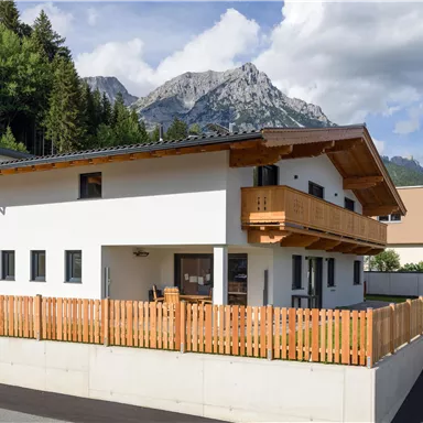 Ein modernes Haus mit einem Holzbalkon und einem schönen Garten. Im Hintergrund sind beeindruckende Berge und blauer Himmel zu sehen.