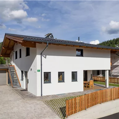 Ein modernes, zweigeschossiges Haus mit einem Holzdach und weißen Wänden. Der Außenbereich ist mit einer Terrasse und einem Holzzaun gestaltet.