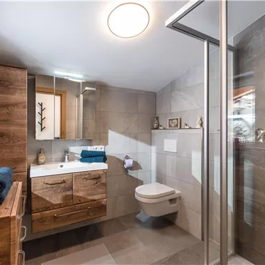 Ein modernes Badezimmer mit Holzdetails und modernen Fliesen. Es verfügt über eine Dusche, eine Toilette und ein Waschbecken.