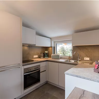 Eine moderne Küche mit weißen Schränken und einer grauen Arbeitsplatte. Das Zimmer hat Fenster, die Licht hereinlassen.