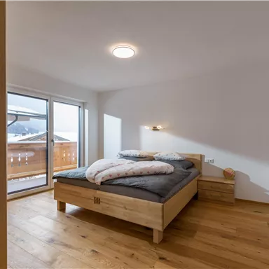 Ein helles Schlafzimmer mit einem Holzbett und zwei Kissen. Große Fenster bieten einen Blick auf eine Terrasse.